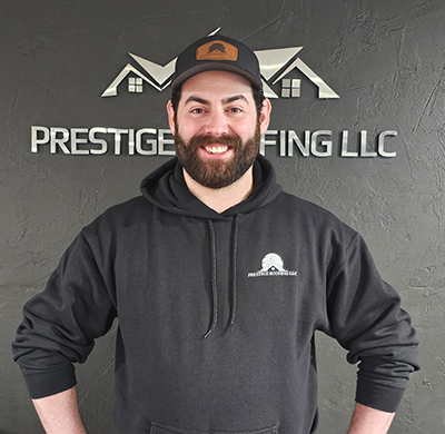 Smiling man in black hoodie in front of Prestige Roofing LLC sign.